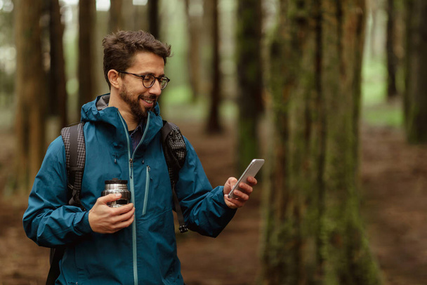 Positive young caucasian couple in jackets with cup of coffee, walk in forest together, enjoy trip, use phone map, outdoor. Hiking, lifestyle, adventure and tourism alone with device - Photo, Image