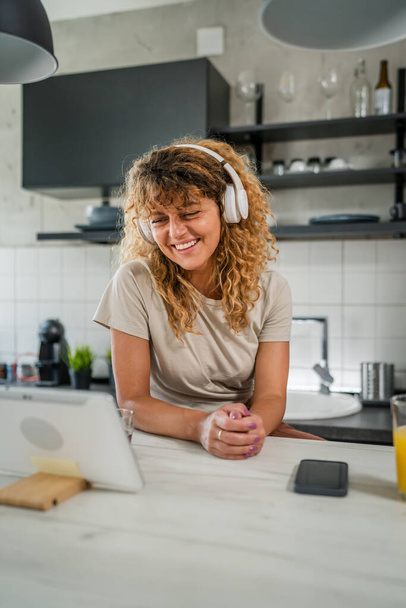 Uma mulher caucasiana com cabelo encaracolado sentar em casa uso tablet digital e fones de ouvido para assistir filme ou série de fluxo on-line ou para ter chamada de vídeo sorriso feliz ter um bom espaço de cópia do tempo - Foto, Imagem