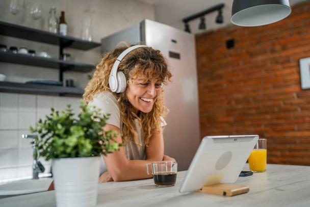 Une femme caucasienne avec les cheveux bouclés assis à la maison utiliser tablette numérique et écouteurs pour regarder un film ou une série en ligne flux ou d'avoir un appel vidéo sourire heureux avoir un bon espace de copie de temps - Photo, image