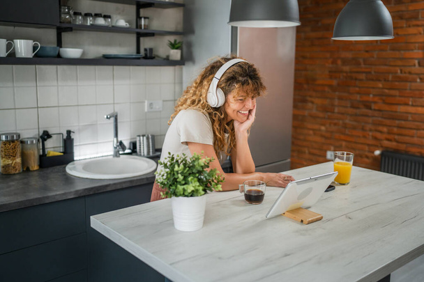 Eine kaukasische Frau mit lockigem Haar sitzt zu Hause und nutzt ein digitales Tablet und Kopfhörer, um Filme oder Serien online zu sehen oder Videoanrufe zu führen. - Foto, Bild