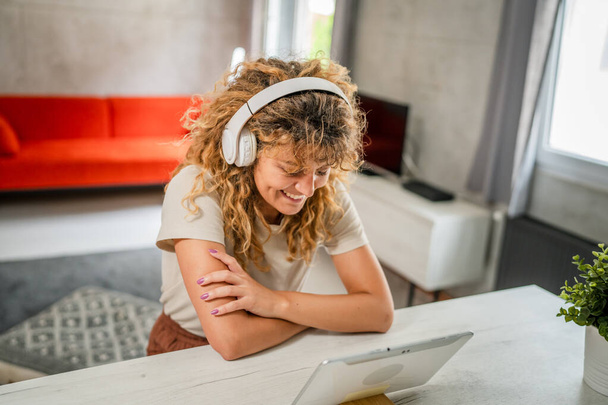 Uma mulher caucasiana com cabelo encaracolado sentar em casa uso tablet digital e fones de ouvido para assistir filme ou série de fluxo on-line ou para ter chamada de vídeo sorriso feliz ter um bom espaço de cópia do tempo - Foto, Imagem