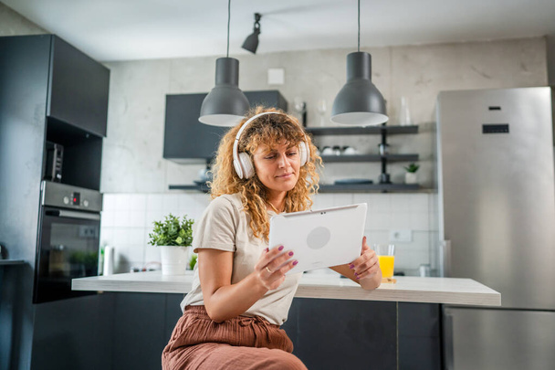 Μια καυκάσια γυναίκα με σγουρά μαλλιά κάθονται στο σπίτι χρησιμοποιούν ψηφιακή ταμπλέτα και ακουστικά για να παρακολουθήσουν ταινία ή σειρά σε απευθείας σύνδεση ρεύμα ή να έχουν κλήση βίντεο χαρούμενο χαμόγελο έχουν μια καλή στιγμή αντίγραφο χώρο - Φωτογραφία, εικόνα