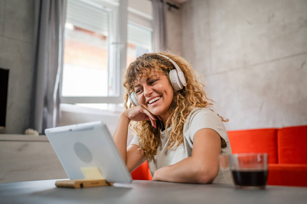 One caucasian woman with curly hair sit at home use digital tablet and headphones to watch movie or series online stream or to have video call happy smile have a good time copy space - Photo, Image