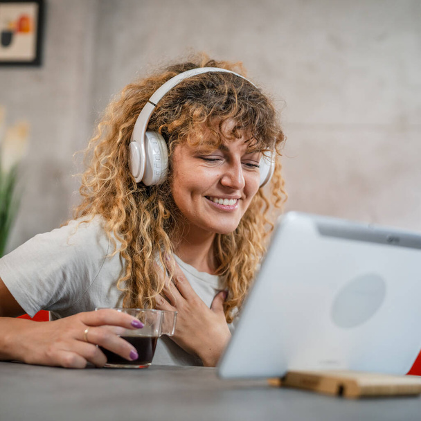 Одна біла жінка з кучерявим волоссям сидить вдома, використовує цифровий планшет та навушники для перегляду фільмів або серіалів онлайн-потоку або для відеодзвінків щаслива посмішка має гарне місце для копіювання - Фото, зображення