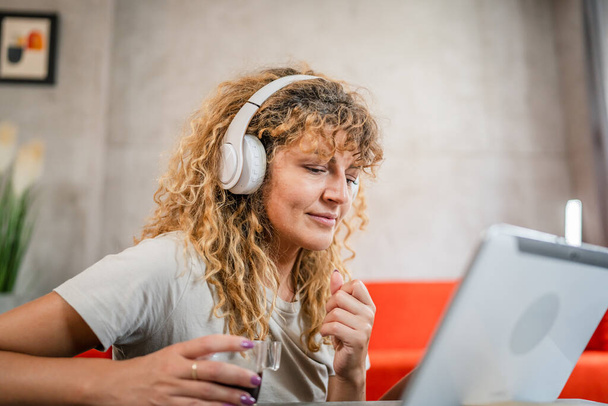 One caucasian woman with curly hair sit at home use digital tablet and headphones to watch movie or series online stream or to have video call happy smile have a good time copy space - Foto, Bild