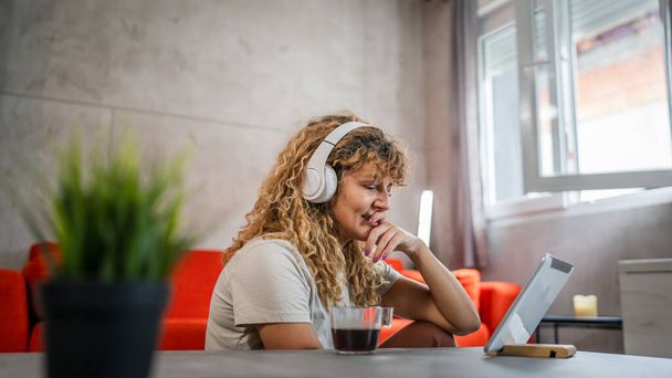 One caucasian woman with curly hair sit at home use digital tablet and headphones to watch movie or series online stream or to have video call happy smile have a good time copy space - Photo, Image