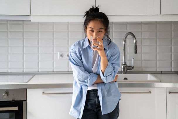 Unglücklich zerzaustes asiatisches Mädchen trinkt Wasser aus Glas, während es am späten Morgen in der Küche steht und schlechte Laune weckt und ein Antidepressivum trinkt. Koreanische kranke Frau mittleren Alters verdauungsgestört und durstig. - Foto, Bild