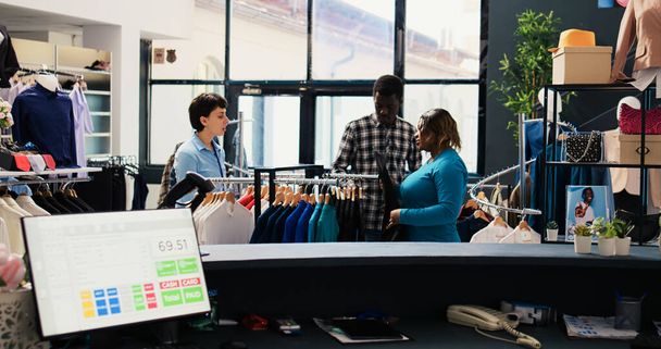 Υπάλληλος καταστήματος βοηθώντας ζευγάρι με μοντέρνα ρούχα, δείχνοντας κρεμάστρες με νέα συλλογή μόδας στη σύγχρονη μπουτίκ. Αφροαμερικανοί πελάτες που ψωνίζουν για κομψά εμπορεύματα στο κέντρο ένδυσης - Φωτογραφία, εικόνα