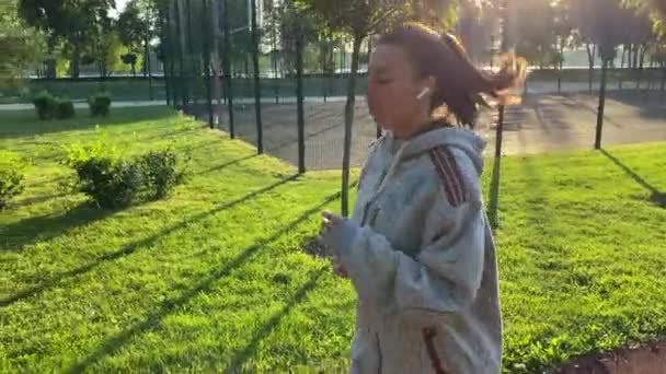 Side view multiracial young woman jogging on running trail in golden light of rising sun. Female jogger using wireless earbuds exercising in public park. Healthy wellbeing, urban fitness lifestyle - Footage, Video