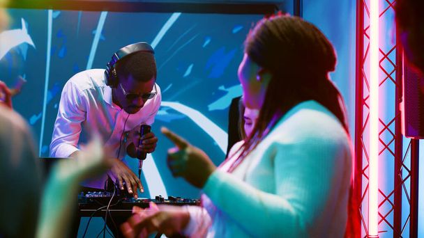 クラブで楽しんでいるアフリカ系アメリカ人DJ 、ディスコでオーディオステーションで電子音楽をミックスします。若い男はステージ上の人々と幸せなパーティーを感じます,ナイトクラブでサウンドパネルを使用して. - 写真・画像