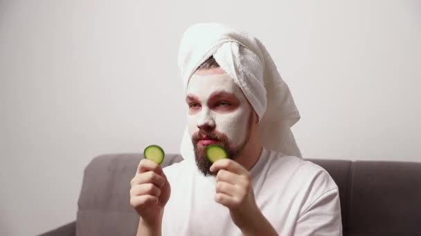 Πορτρέτο του αστείου άνδρα σε λευκό μπλουζάκι εφαρμόζοντας φέτες αγγούρι πάνω από πηλό μάσκα στο πρόσωπό του. Πρωινή διαδικασία σπα. Έννοια επεξεργασίας ομορφιάς - Πλάνα, βίντεο