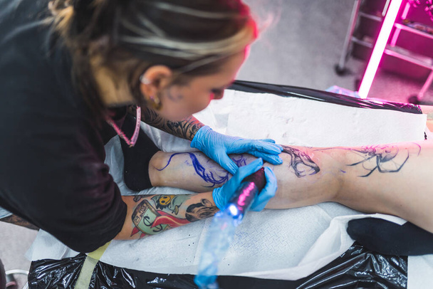 Tatuaggio artista donna che disegna un tatuaggio su una gamba clienti. Concetto art. Alto angolo di tiro al coperto. Sfondo sfocato. Foto di alta qualità - Foto, immagini