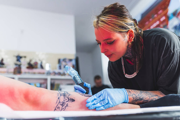 Tattooing at art studio. Girl tattoo artist makes tattoo on a leg. Blurred background. Focusing on ones job. High quality photo - Photo, Image