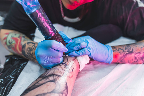 Крупный план профессионального процесса татуировки с использованием гигиенических продуктов, таких как латексные перчатки. Блестящая раздражённая кожа делает татуировку. Высокое качество фото - Фото, изображение