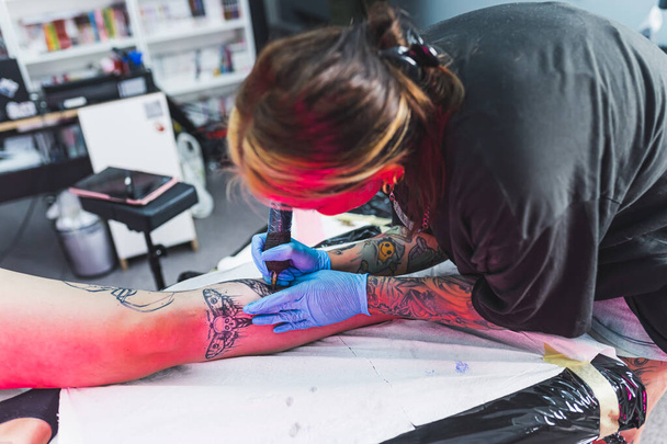 Sanat becerisi gerektiren bir iş. Müşterisinin bacağına dövme çizen odaklanmış bir kadın sanatçı. Arka plan bulanık. Dövme oturumunda dövmeli yatakta yatan bir müşteri. Yüksek kalite fotoğraf - Fotoğraf, Görsel