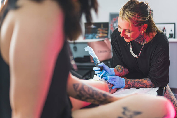 Muotokuva nauravasta onnellisesta naisesta tatuointipäälliköstä, joka näyttää luomisprosessin tatuoinnin jalassa. Positiivinen työympäristö. Laadukas kuva - Valokuva, kuva