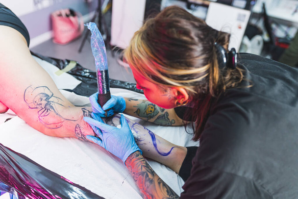 Сфокусированная девушка-татуировщица наклоняется, чтобы нарисовать точную татуировку на ноге своего первого клиента. Выстрел под высоким углом. Высокое качество фото - Фото, изображение