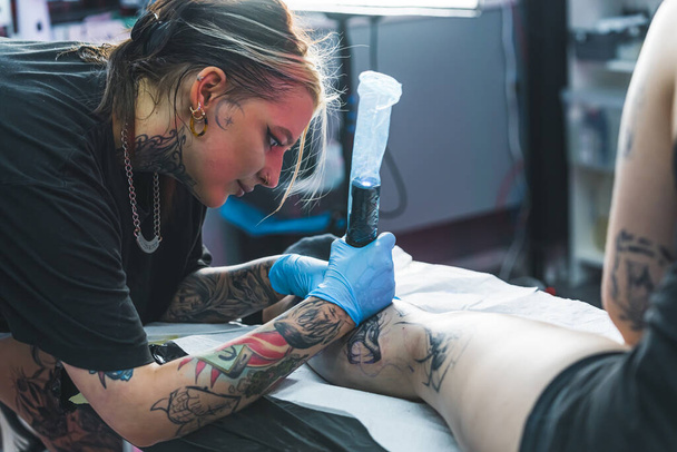 Mladý umělec během tetování. Tetovaná umělkyně kreslí tetování nohy. Umění v mnoha podobách. Vnitřní záběr. Kvalitní fotografie - Fotografie, Obrázek