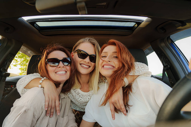 Bonnes jeunes femmes amies dans des vêtements décontractés à la mode et des lunettes de soleil embrassant en groupe tout en étant assis dans la voiture souriant et regardant la caméra - Photo, image
