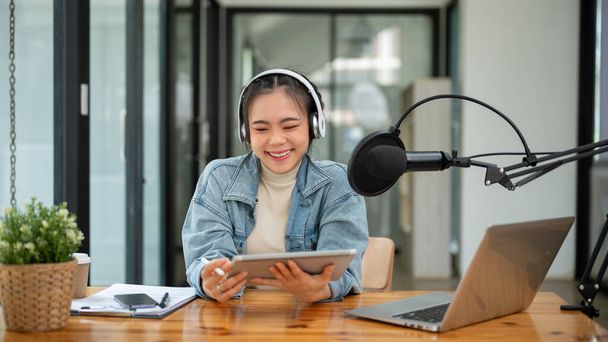 Piękna i wesoła Azjatka prowadząca radio lub podcaster patrzy na ekran tabletu, opowiada historię, przemawia do mikrofonu i nagrywa swój podcast w swoim studio. - Zdjęcie, obraz