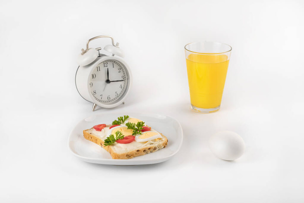Тарелка с бутербродом с яйцом, помидорами и петрушкой на фоне часов. Рядом со стаканом сока. Концепция здорового питания. Пространство для текста - Фото, изображение
