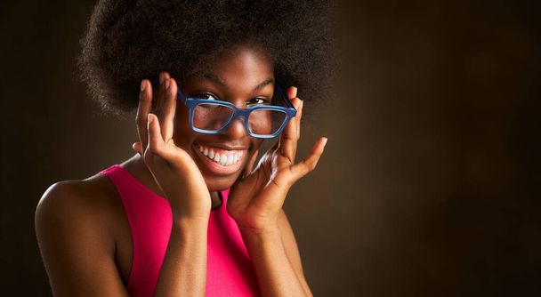 молодая этническая женщина с африканской прической счастливо улыбается, глядя в камеру в синих очках и розовой футболке - Фото, изображение