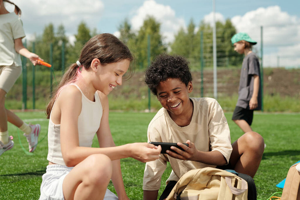 Двое веселых школьников смеются над смешным онлайн видео в мобильном телефоне, сидя на зеленом футбольном поле или стадионе перед камерой - Фото, изображение
