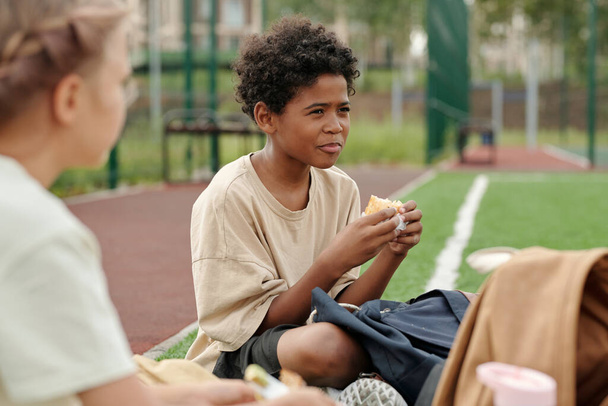 Cute głodny Afroamerykanin chłopiec w casualwear jedzenia kanapki lub inne przekąski na lunch podczas siedzenia wśród kolegów z klasy lub przyjaciół - Zdjęcie, obraz