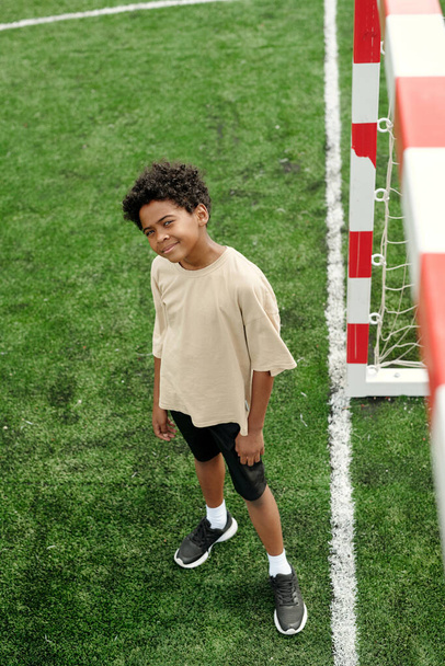 Por encima del ángulo de lindo colegial afroamericano en ropa deportiva mirando a la cámara mientras está de pie en el campo de fútbol verde en el estadio en el ocio - Foto, Imagen