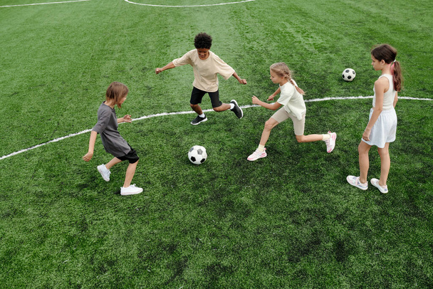 サッカーボールに続く異文化間の子供たちのグループは,ゲーム中に緑のサッカー場に沿って走っているか,物理的なトレーニングのレッスン - 写真・画像