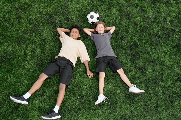 Vysoký úhel dvou klidných mezikulturních kluků v aktivním oblečení ležících na zeleném trávníku nebo fotbalovém hřišti, zatímco si užívají přestávku po hraní fotbalu - Fotografie, Obrázek