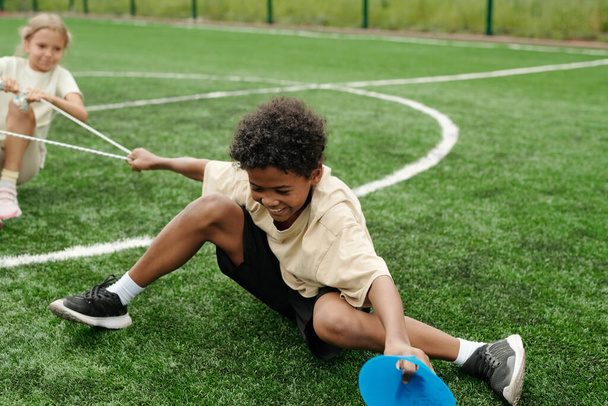 Vrolijke Afro-Amerikaanse schooljongen trekken touw en weerstand tegen klasgenoot tijdens sportcompetitie op stadion of voetbalveld - Foto, afbeelding