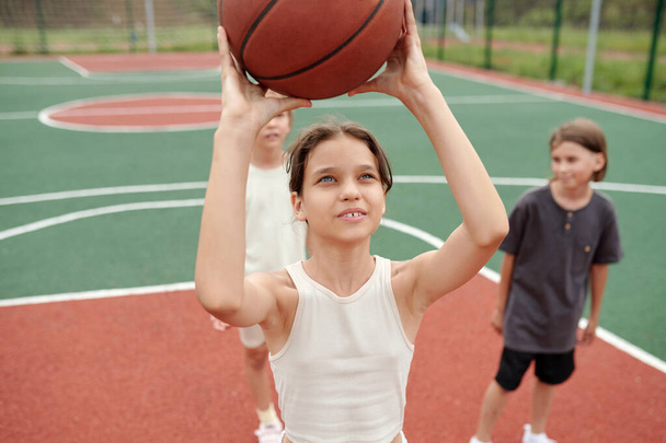Cute schoolgirl in activewear throwing ball into basket while standing in front of camera against classmates waiting for their turn to play - Photo, Image