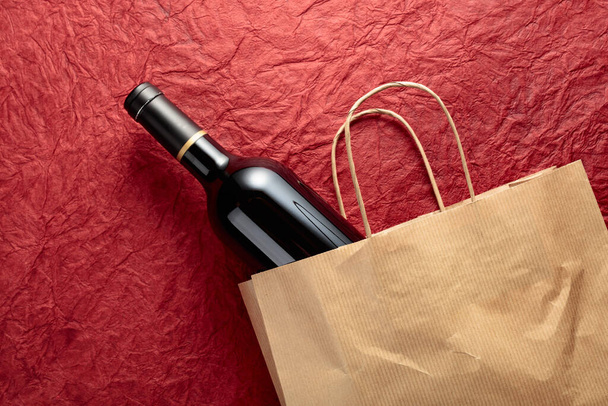 質感のある赤の背景に赤ワインのボトルと紙のショッピングバッグ。トップビュー、コピースペース. - 写真・画像