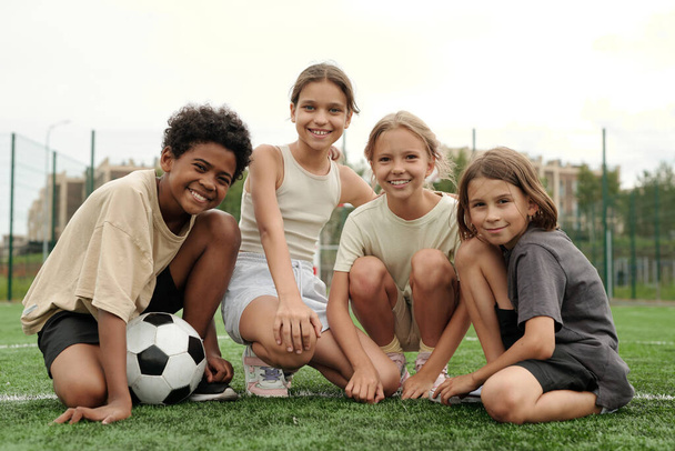 Team aus vier aktiven Freunden und Fußballern schaut lächelnd in die Kamera, während sie auf dem grünen Rasen des Fußballfeldes am Stadion sitzen - Foto, Bild