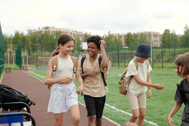 Dwoje uroczych międzykulturowych uczniów rozmawia o czymś podczas rozmowy, spacerując po przestronnym stadionie z kolegami z klasy i śmiejąc się - Zdjęcie, obraz