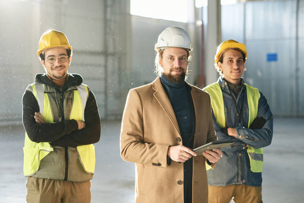 Onnellinen nuori työnjohtaja beige takki ja kaksi monietninen insinöörit suojakypärät ja turvallisuus heijastava takit seisoo kameran edessä - Valokuva, kuva