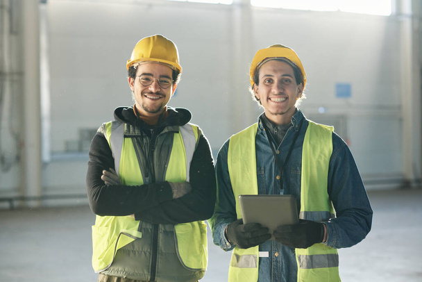 工場や工場の倉庫でカメラの前に立っているハードハットと反射ジャケットの2人の幸せな若いマルチエスニックエンジニア - 写真・画像