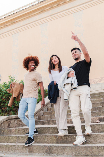 Retrato vertical de un grupo de estudiantes multiétnicos caminando juntos abajo del campus, hablando, señalando y sonriendo. Joven adolescente mujer con dos amigos masculinos turismo en un viaje - Foto, Imagen