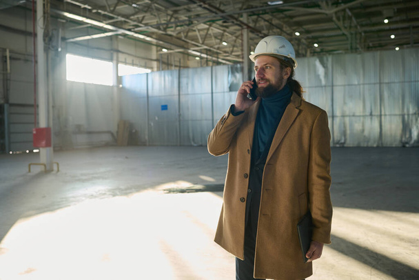 Nuori menestyvä insinööri casualwear ja kypärä puhuu matkapuhelimella työpajassa keskustellessaan työpisteistä alaistensa kanssa - Valokuva, kuva