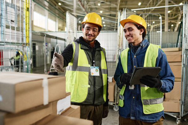 Счастливый молодой человек в рабочей одежде с планшетом и слушая, как коллега указывает на стопку упакованных коробок на металлической тележке во время работы - Фото, изображение