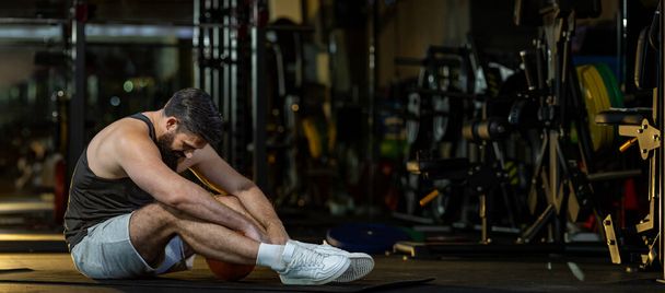Homme caucasien est confronté au syndrome de surentraînement après l'entraînement à l'intérieur de la salle de gym avec fond sombre pour l'exercice et de remise en forme - Photo, image