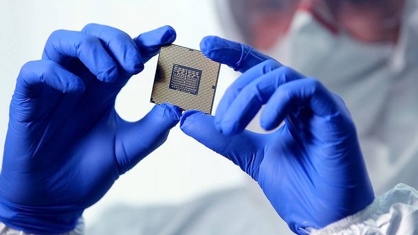 Hochmoderne Fabrik für elektronische Fertigung, Ingenieur im sterilen Overall hält Mikrochip mit Handschuhen und untersucht ihn - Foto, Bild