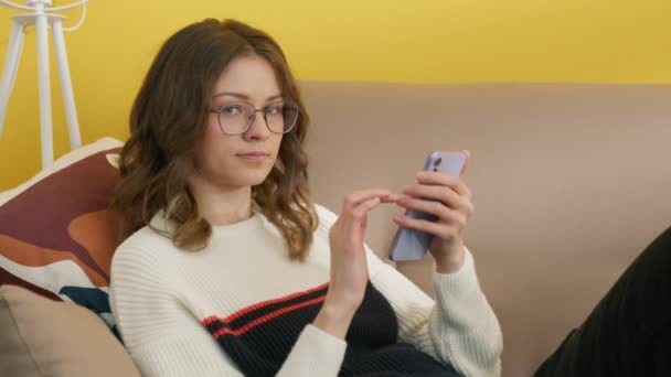 Εστιασμένη κοπέλα κύλιση οθόνη αφής στο smartphone. Γυναίκα φοιτήτρια ξεκουράζεται στον καναπέ μετά από online μαθήματα. Χαρούμενη γυναίκα με γυαλιά που κοιτάει την κάμερα. Νεαρή κοπέλα που χρησιμοποιεί κινητό τηλέφωνο στο σπίτι, 4k πλάνα - Πλάνα, βίντεο