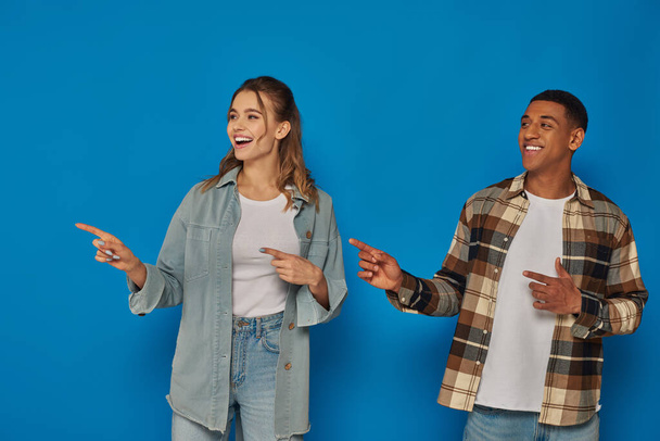 happy woman with open mouth and african american man pointing away with fingers on blue background - Foto, Imagem