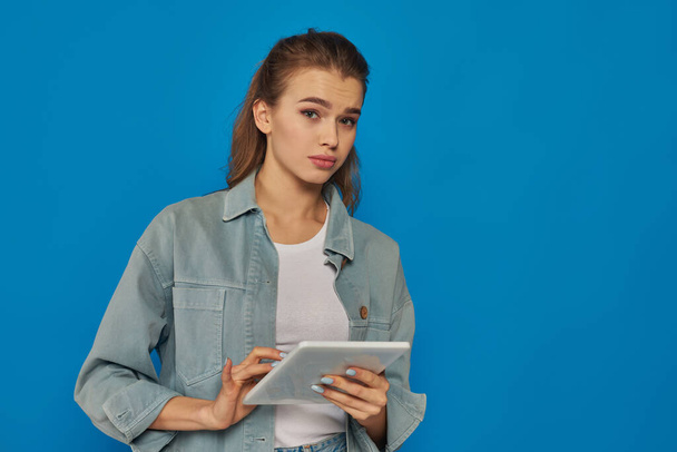 όμορφη νεαρή γυναίκα κρατώντας ψηφιακό tablet και κοιτάζοντας κάμερα σε μπλε φόντο, σύγχυση πρόσωπο - Φωτογραφία, εικόνα