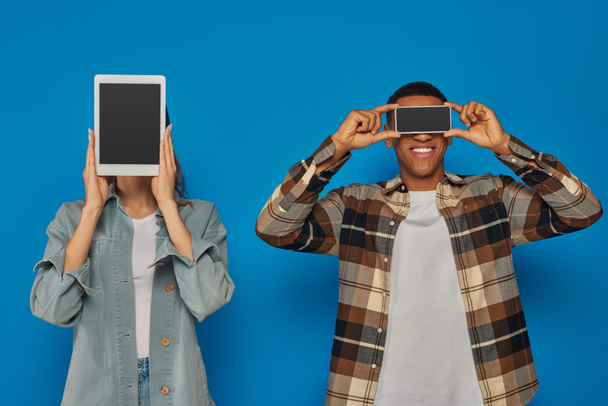 pareja interracial celebración de teléfono inteligente y tableta digital con pantalla en blanco sobre fondo azul - Foto, imagen