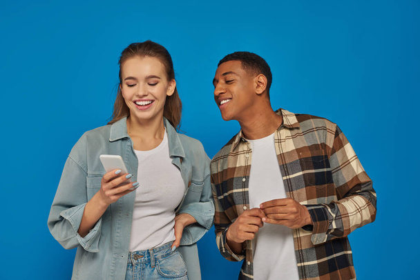 szczęśliwy african amerykański mężczyzna patrząc na smartfon z kobieta przyjaciel i uśmiechając się na niebieskim tle - Zdjęcie, obraz