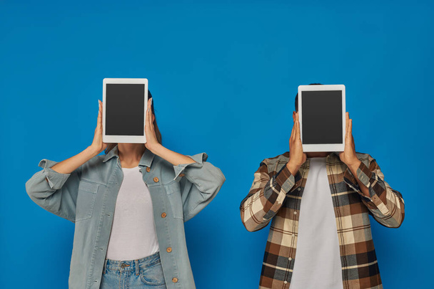 interracial homme et femme obscurcissant visage avec des tablettes numériques sur fond bleu, écran tactile vierge - Photo, image