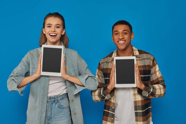 hombre y mujer interracial feliz sosteniendo tabletas digitales con pantalla en blanco sobre fondo azul - Foto, Imagen